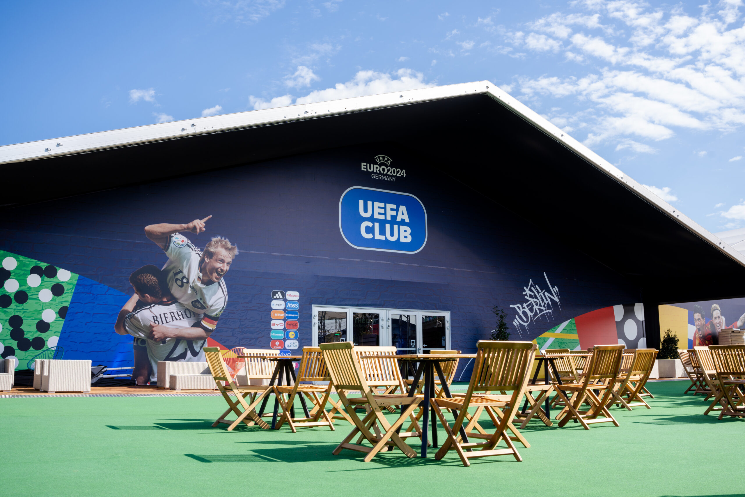 Außenansicht des UEFA Club Gebäudes mit dunkler Textilhülle und Holzbestuhlung im Außenbereich